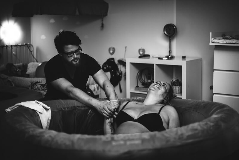 Frau im Geburtspool, Partner hält ihre Hand, Geburtsfotografie